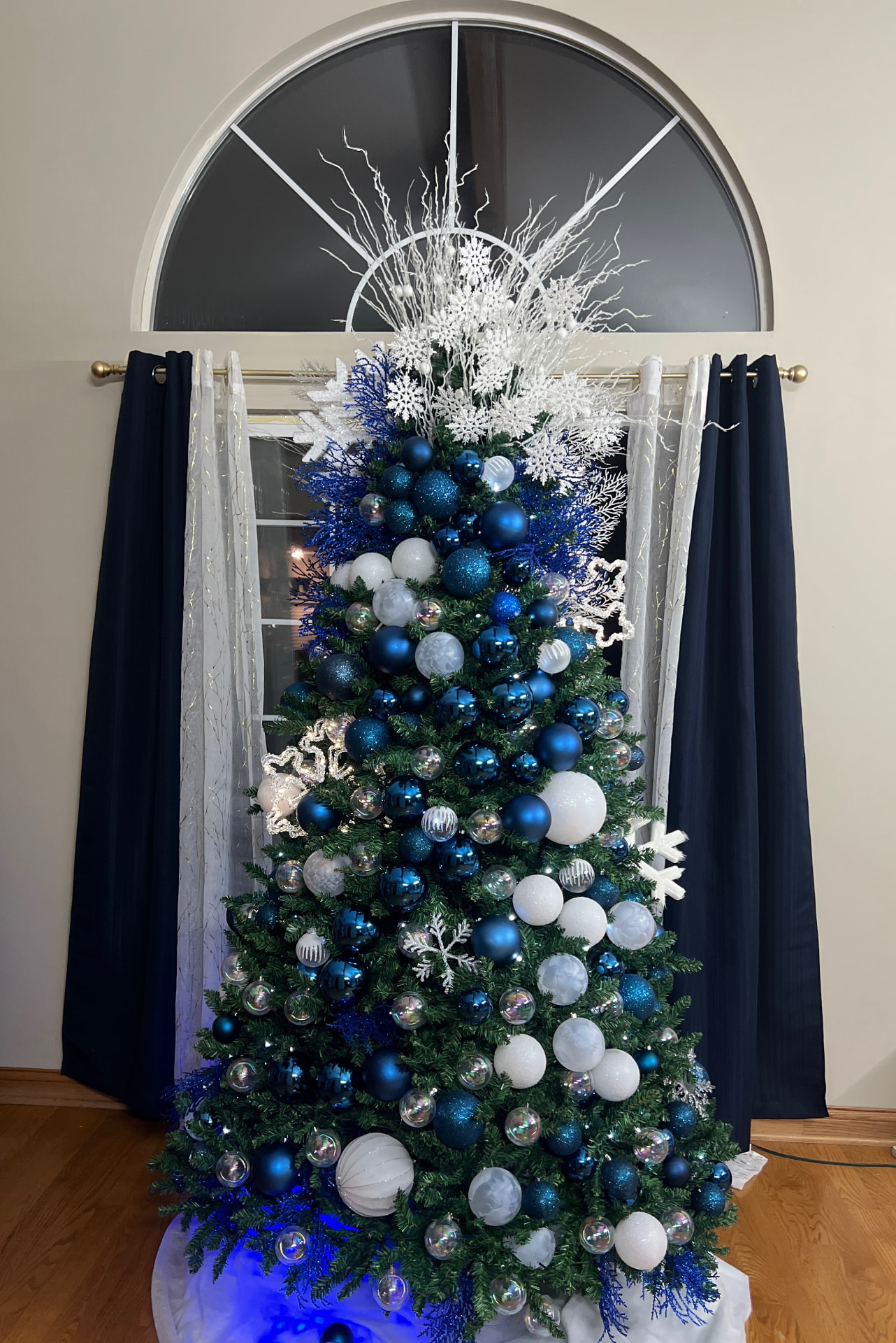 Blue and white winter-themed Christmas tree with snowflake picks in front of an arched window with navy curtains