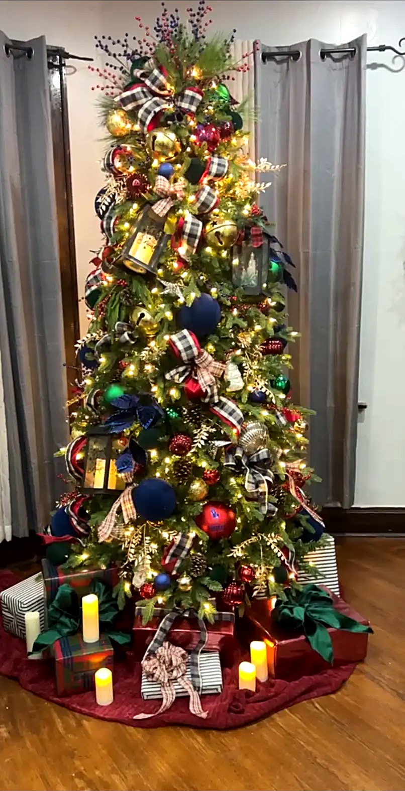 Rustic Christmas tree with buffalo plaid ribbon, glowing lanterns, navy and red ornaments, candles, and wrapped presents